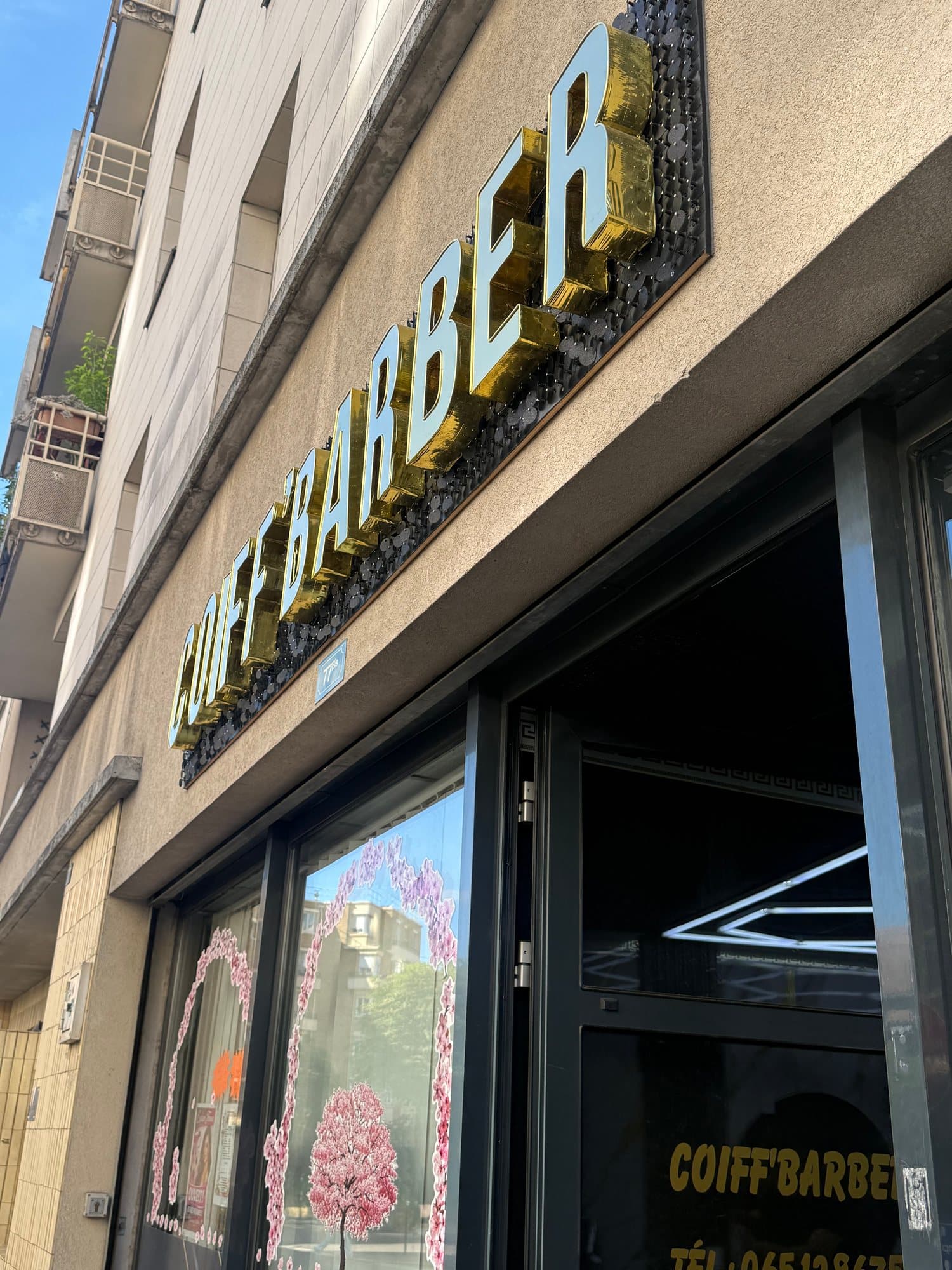 L'enseigne dorée COIFF'BARBER vue d'en bas, au dessus de la vitrine fleurie