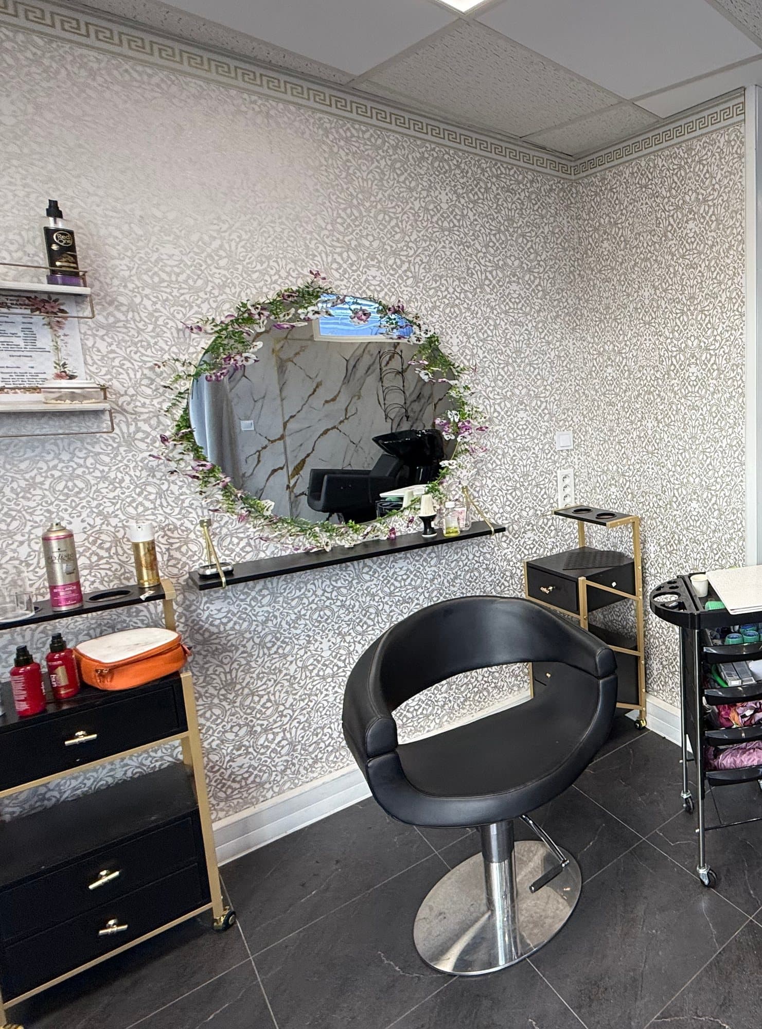 Espace Femme : fauteuil de coiffure face à un miroir rond couronné de fleurs, mur en papier peint argenté